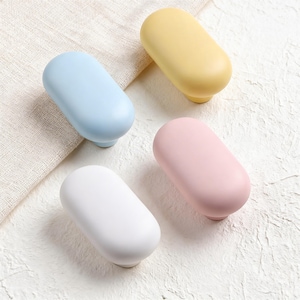 Può includere: Quattro pomelli ovali per armadi in colori pastello: azzurro, giallo, rosa e bianco. Ogni pomello ha una parte superiore arrotondata e una base cilindrica corta. I pomelli sono disposti su una superficie bianca strutturata.