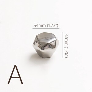 Op de afbeelding: Een gepolijste zilverkleurige kastknop met een facet, geometrisch ontwerp. De knop meet 44 mm breed en 32 mm hoog. De letter "A" is in bruin gedrukt in de linker benedenhoek.