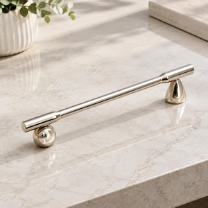 May include: A polished nickel cabinet pull featuring a long, slender horizontal bar supported by two distinct bases. One base is a spherical shape, while the other is a tapered, conical form. The metal has a reflective, smooth, and metallic finish.