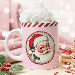 May include: A pink ceramic mug with a Santa Claus illustration, filled with whipped cream. The mug has a white handle and a red and white striped rim. Holly and berries are in the foreground, with cookies in the background.