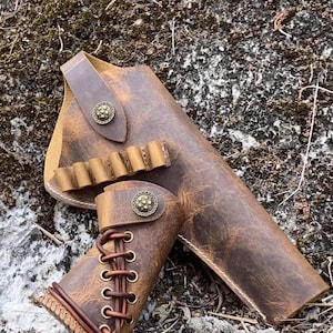 以下が含まれることがあります： 手作りの茶色の革製ガンホルスターとカートリッジホルダーのセット。革は使い古したような仕上がりで、装飾的な金属ボタンのアクセントが付いています。ホルスターはレースのデザインで、レースの端には真鍮色の金属ボールが付いています。