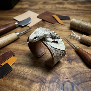 Peut inclure: Un bracelet manchette en cuir artisanal avec un motif de serpent. La tête du serpent est blanche et noire, avec une langue fourchue. Le bracelet est marron et repose sur une surface en bois, entouré d'outils de maroquinerie.