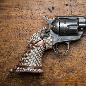 Könnte beinhalten: Ein Vintage-Revolver mit einem individuellen Schlangenhautgriff, braunen Lederschnürsenkeln und einem bronzenen Schlangenemblem. Die Waffe befindet sich vor einem hölzernen Hintergrund.