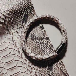 May include: A white and gray snake skin bracelet with a black magnetic clasp. The bracelet is circular and rests on a background of snake skin. The scales are white, gray, and black.