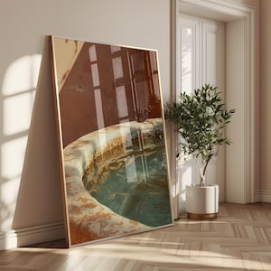 Puede incluir: Una impresión artística enmarcada que presenta un primer plano de una fuente circular y desgastada llena de agua. El agua tiene un tono turquesa. El marco es dorado y el fondo muestra una pared con reflejos de ventanas. Una planta en maceta está al lado del marco.