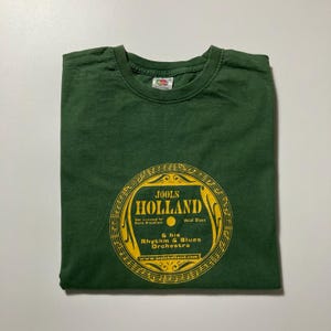 Könnte beinhalten: Dunkelgrünes T-Shirt mit gelbem, kreisförmigem Aufdruck mit der Aufschrift "JOOLS HOLLAND" und "& his Rhythm & Blues Orchestra". Der Aufdruck enthält auch die Website-Adresse www.joolsholland.com. Das Shirt ist aus weichem Material.
