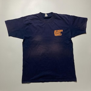Puede incluir: Camiseta azul marino de manga corta. El texto naranja "JOE COCKER RESPECT YOURSELF" está impreso en la parte superior derecha del pecho. Cuello redondo, la camiseta parece vintage.