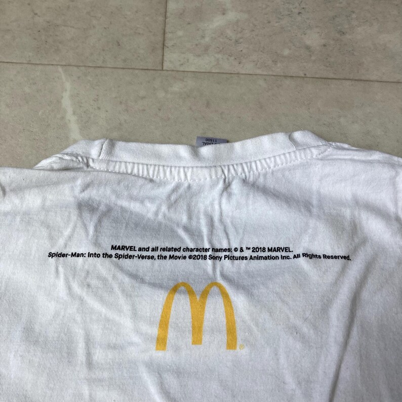 K&ouml;nnte beinhalten: Ein wei&szlig;es T-Shirt mit einem gelben McDonald's-Logo in Form goldener B&ouml;gen, gedruckt unter einem schwarzen Text, der lautet: MARVEL and all related character names: &copy; & &trade; 2018 MARVEL. Spider-Man: Into the Spider-Verse, the Movie &copy;2018 Sony Pictures Animation Inc. All Rights Reserved.