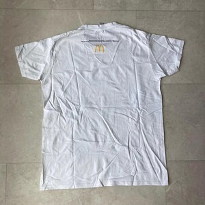 K&ouml;nnte beinhalten: Ein wei&szlig;es kurz&auml;rmeliges T-Shirt mit einem kleinen gelben McDonald's-Logo in Form goldener B&ouml;gen, das mittig nahe dem Halsausschnitt platziert ist. Das Shirt hat einen Rundhalsausschnitt und eine schlichte R&uuml;ckseite mit kleinem schwarzem Text.