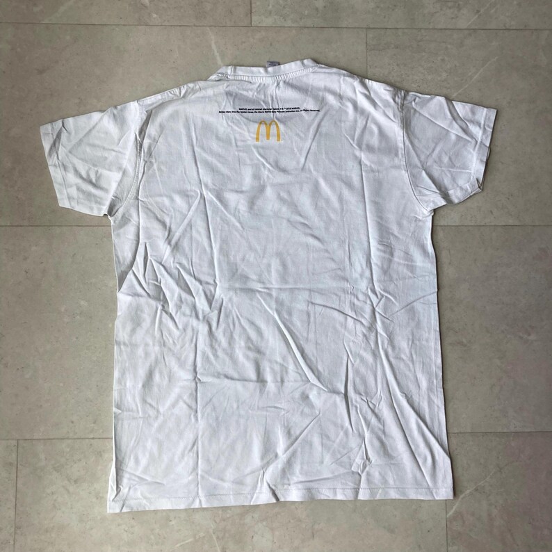 K&ouml;nnte beinhalten: Ein wei&szlig;es kurz&auml;rmeliges T-Shirt mit einem kleinen gelben McDonald's-Logo in Form goldener B&ouml;gen, das mittig nahe dem Halsausschnitt platziert ist. Das Shirt hat einen Rundhalsausschnitt und eine schlichte R&uuml;ckseite mit kleinem schwarzem Text.