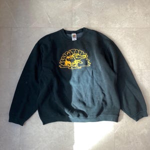 Könnte beinhalten: Dunkelgrünes Sweatshirt mit Rundhalsausschnitt und langen Ärmeln. Auf der Vorderseite befindet sich eine gelbe Grafik eines bärenartigen Wesens mit dem Text "BIG TOP". Das Sweatshirt hat eine klassische, lockere Passform.