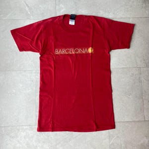 May include: A short-sleeved red t-shirt featuring the word BARCELONA printed in white capital letters across the chest. The text is underlined by thin horizontal lines and accompanied by a small yellow circular graphic on the right side.