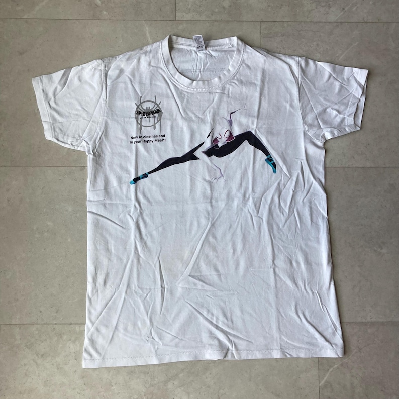 K&ouml;nnte beinhalten: Ein wei&szlig;es Kurzarm-T-Shirt mit einer Grafik von Spider-Gwen in einer dynamischen Pose. Das Shirt enth&auml;lt das Spider-Man Across the Spider-Verse Logo und den Text: Now in cinemas and in your Happy Meal!