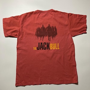 Op de afbeelding: Rood t-shirt met een afbeelding van silhouetten en de tekst "THE JACK BULL" in zwart en geel. Het shirt heeft korte mouwen en een ronde hals.