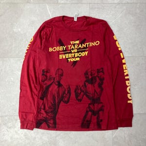 Puede incluir: Camiseta roja de manga larga con texto amarillo "THE BOBBY TARANTINO VS EVERYBODY TOUR". La camiseta presenta ilustraciones negras de dos figuras. La manga derecha tiene la palabra "EVERYBODY" en amarillo.