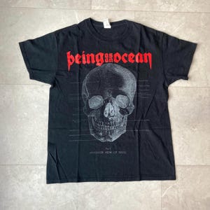 May include: Black t-shirt featuring a skull graphic with the text "being as ocean" in red. The skull is a detailed anatomical illustration with the words "anterior view of skull" below it. The shirt is a crew neck.