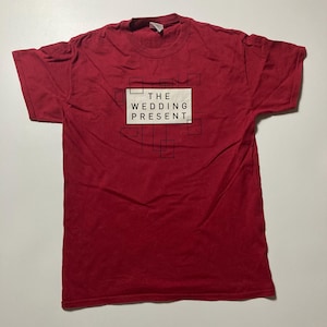 Może przedstawiać: Czerwona koszulka z napisem "THE WEDDING PRESENT" w białym prostokącie, otoczonym czarnymi geometrycznymi kształtami. Koszulka jest w jednolitym czerwonym kolorze i wydaje się być wykonana z bawełny.
