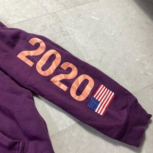 May include: A purple hoodie sleeve with the number "2020" in a peach color printed on the sleeve. An American flag patch is also visible on the sleeve.