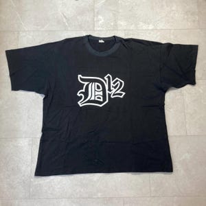 May include: A black short-sleeved t-shirt featuring a large white graphic print on the chest. The graphic displays the stylized text D12 in a bold, gothic-inspired font with a white outline, centered against the solid black fabric of the shirt.