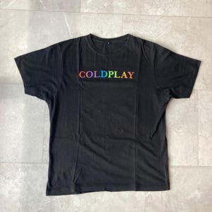 Puede incluir: Camiseta negra con la palabra "COLDPLAY" en letras de colores del arcoíris en el pecho. La camiseta de manga corta tiene cuello redondo. La tela parece ser de algodón suave.