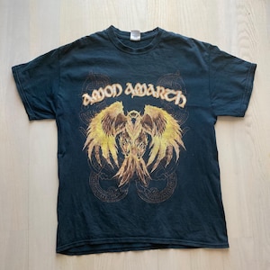 Puede incluir: Camiseta de color verde azulado oscuro con el nombre de la banda "Amon Amarth" sobre un gráfico detallado de un pájaro con las alas extendidas en tonos amarillos y marrones. El diseño incluye patrones de inspiración vikinga.