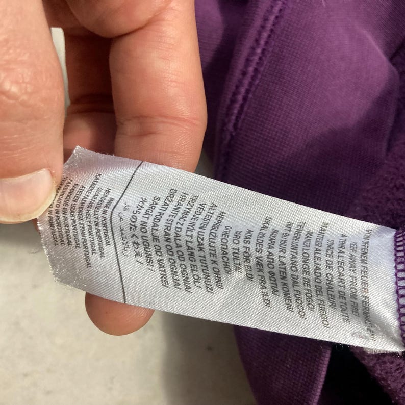 May include: A close-up of a purple fabric with a white tag attached. The tag has text in multiple languages, including English, German, and Japanese. The tag reads "Made in Portugal" and includes fire safety warnings.