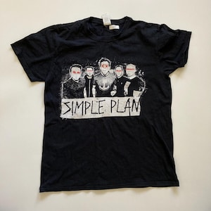 May include: Black t-shirt with a graphic of a band with red accents over their eyes and the band name "SIMPLE PLAN" in white block letters. The shirt is made of a soft, dark fabric.