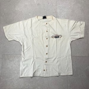 May include: A cream-colored short-sleeve button-down shirt featuring a Central Perk logo on the left chest. The shirt has a round neckline, a vertical row of brown buttons down the center, and a small black tag at the inner collar area.