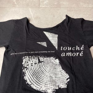 May include: Black t-shirt with white graphic designs and text. The shirt features a large, textured, circular design, geometric shapes, and the words "touch&eacute; amor&eacute;." Additional text reads "give me a reason not to just start screaming out loud."