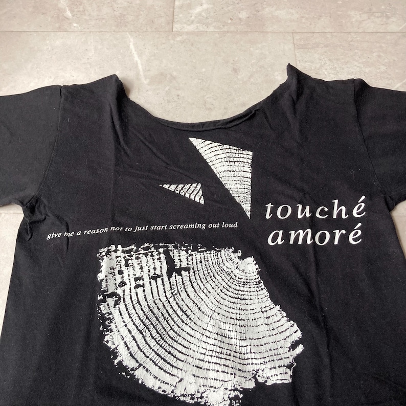 May include: Black t-shirt with white graphic designs and text. The shirt features a large, textured, circular design, geometric shapes, and the words "touch&eacute; amor&eacute;." Additional text reads "give me a reason not to just start screaming out loud."
