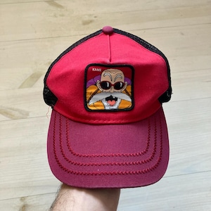 Peut inclure: Casquette de camionneur rouge et noir avec un écusson de personnage de dessin animé portant des lunettes de soleil et le texte "KAME". La casquette a une visière incurvée et un dos en filet.