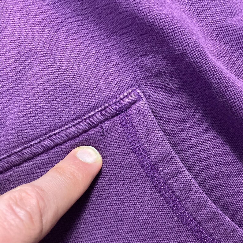 May include: Close-up of a section of a purple fabric, possibly a garment. The fabric has a textured appearance with visible stitching along the edges. A finger is pointing at a small imperfection in the fabric.