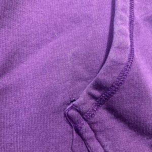 May include: Close-up of a section of a purple garment, possibly a sweatshirt or hoodie. The fabric has a ribbed texture, with visible stitching along the seams. The color is a vibrant, solid purple.