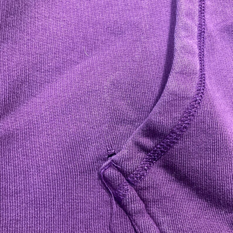 May include: Close-up of a section of a purple garment, possibly a sweatshirt or hoodie. The fabric has a ribbed texture, with visible stitching along the seams. The color is a vibrant, solid purple.