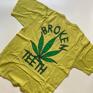 Può includere: T-shirt giallo con una grande grafica a forma di foglia di cannabis verde. Le parole "BROKEN TEETH" sono stampate in verde sopra e sotto la foglia. La maglietta è realizzata in un materiale morbido.