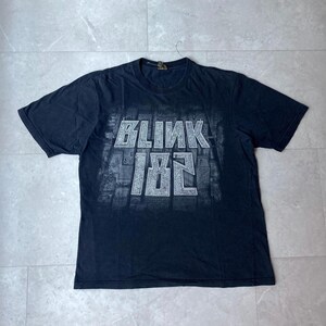 May include: A black short-sleeve t-shirt featuring a white graphic print on the chest. The graphic displays the text "BLINK 182" in a bold, blocky font layered over a detailed, monochromatic illustration of a city skyline grid pattern.