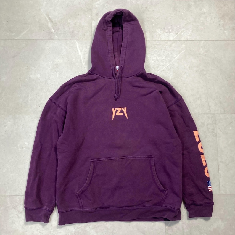 May include: A purple hoodie with a front pocket and a drawstring hood. The front features the letters "YZY" in a peach color. The right sleeve has the word "FORCE" in peach, with a small American flag at the bottom.