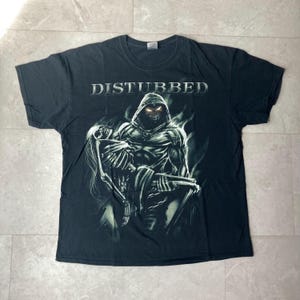 May include: Black t-shirt featuring the band name "DISTURBED" above a graphic of a hooded figure holding a skeleton. The figure has glowing orange eyes and is surrounded by a smoky, ethereal design.