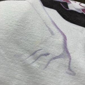 K&ouml;nnte beinhalten: Ein wei&szlig;es T-Shirt mit schwarzem Rippkragen und einem violetten Grafikdruck einer Figur in Yoga-Pose. Der Stoff hat eine sichtbare Strickstruktur und das Design zeigt den stilisierten Umriss einer Person, die sich mit ausgestreckten Gliedma&szlig;en dehnt.
