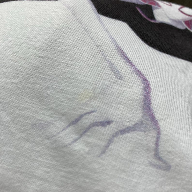 K&ouml;nnte beinhalten: Ein wei&szlig;es T-Shirt mit schwarzem Rippkragen und einem violetten Grafikdruck einer Figur in Yoga-Pose. Der Stoff hat eine sichtbare Strickstruktur und das Design zeigt den stilisierten Umriss einer Person, die sich mit ausgestreckten Gliedma&szlig;en dehnt.