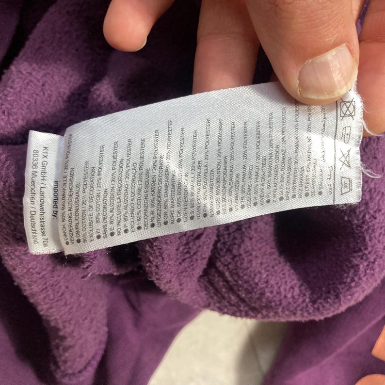 May include: A close-up of a purple garment with a white tag attached. The tag displays text in multiple languages, including English, and details the fabric composition as 80% cotton and 20% polyester. The garment's texture appears soft.
