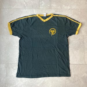 Może przedstawiać: Ciemnoszary t-shirt z krótkim rękawem i dekoltem w serek, wykończony żółtymi lamówkami przy kołnierzyku i rękawach. Na lewej piersi znajduje się żółte, okrągłe logo z literami FF zapisanymi stylizowaną czcionką na materiale.