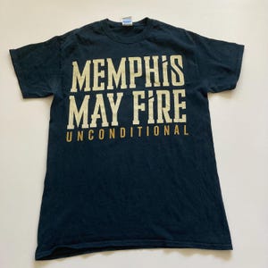 May include: A navy blue t-shirt with the text "MEMPHIS MAY FIRE UNCONDITIONAL" in a gold, bold font. The shirt is laid flat, showcasing the design prominently. The shirt is made of a soft material.