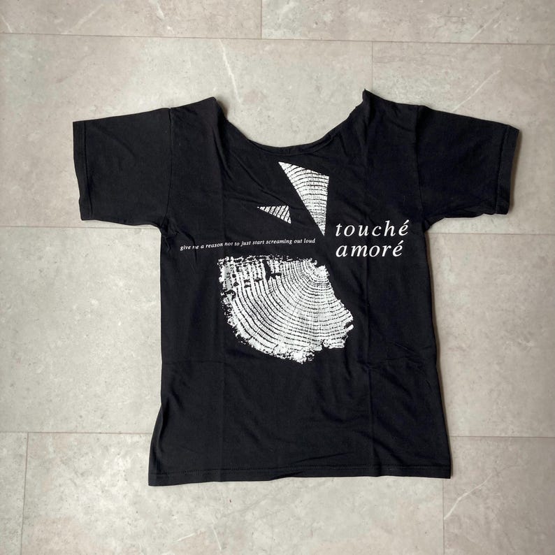 May include: Black t-shirt with a unique neckline and short sleeves. The shirt features white text and graphics, including the words "touch&eacute; amor&eacute;" and a circular design. A line of text is printed below the graphic.