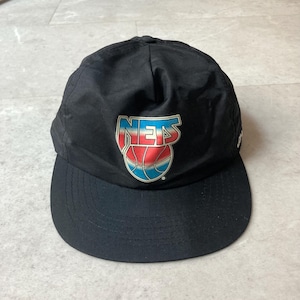 Könnte beinhalten: Eine schwarze Baseballkappe mit einer mittigen Grafik des Wortes NETS in einer rot-weiß-blauen Farbverlaufsschrift über einer Basketball-Illustration in passenden Farben. Die Kappe hat eine strukturierte Krone und einen gebogenen Schirm.