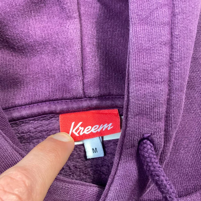 May include: Close-up of a purple hoodie with a drawstring and hood. The red tag reads "Kreem" in white script, with a size "M" tag below. The fabric is a solid, textured material.