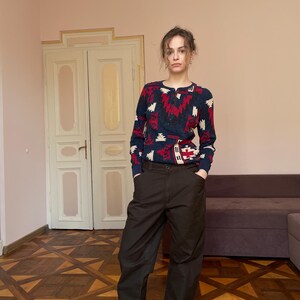 May include: A long-sleeved henley shirt featuring a dark blue base with a repeating geometric pattern in red, white, and black, paired with loose-fitting dark brown trousers and grey slip-on shoes. The shirt has a round neckline with a button placket.