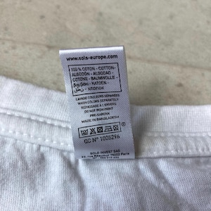 K&ouml;nnte beinhalten: Ein wei&szlig;es Textiletikett an einem wei&szlig;en Kleidungsst&uuml;ck. Das Etikett enth&auml;lt den Text www.sols-europe.com, 100% Baumwolle in mehreren Sprachen, Waschanweisungen, Druck nicht b&uuml;geln, vorgeschrumpft, made in Bangladesh und QC N 1005296.