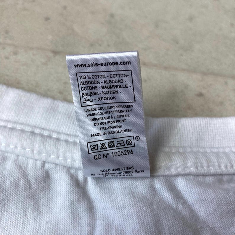 K&ouml;nnte beinhalten: Ein wei&szlig;es Textiletikett an einem wei&szlig;en Kleidungsst&uuml;ck. Das Etikett enth&auml;lt den Text www.sols-europe.com, 100% Baumwolle in mehreren Sprachen, Waschanweisungen, Druck nicht b&uuml;geln, vorgeschrumpft, made in Bangladesh und QC N 1005296.