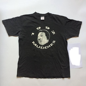 Könnte beinhalten: Schwarzes T-Shirt mit einem weißen Grafikdruck eines Männergesichts, umgeben von dem Text "99N MUDDIES". Das T-Shirt hat einen Vintage-Look und weist leichte Knitterfalten auf.